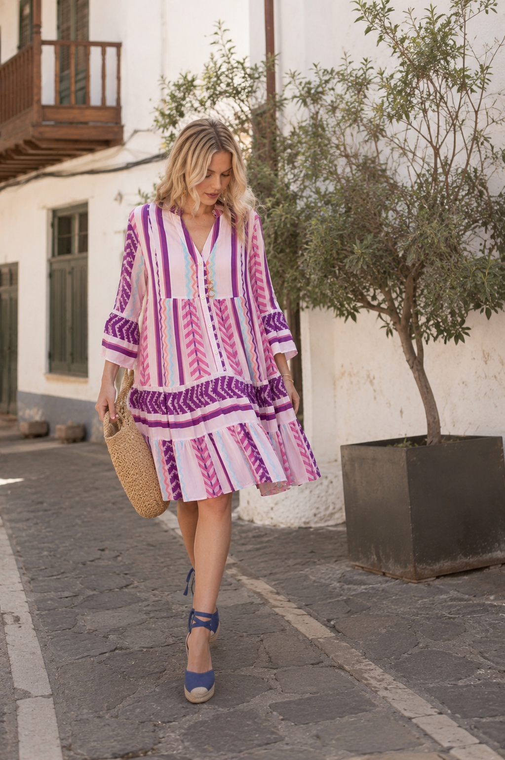 Mujer paseando por un pueblo costero canario con el vestido Dakota estampado en tonos malva, rosa y azul, combinado con alpargatas y bolso de rafia.